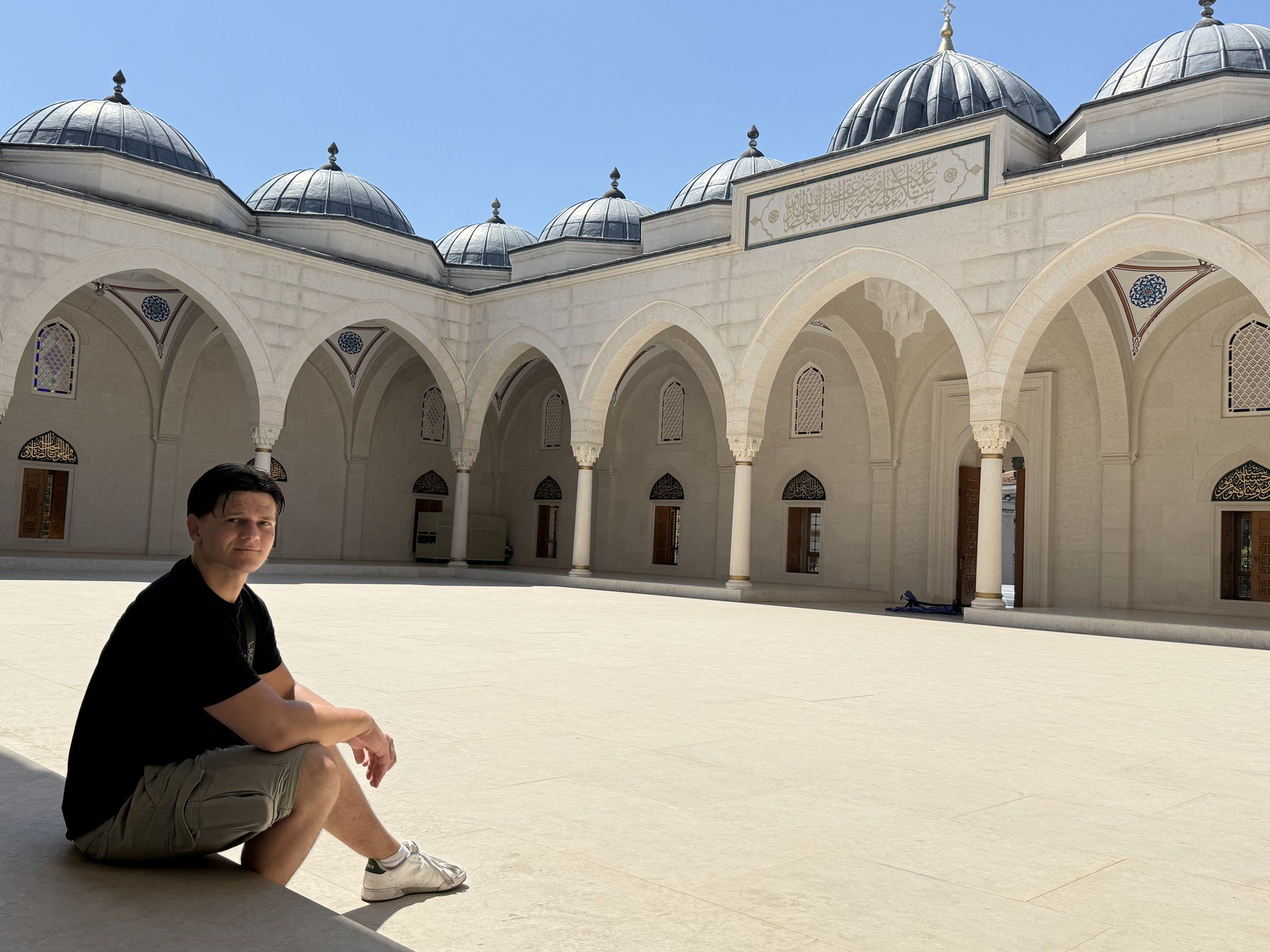 Taras Vasyliv sentado en el patio de piedra clara de una mezquita de Tirana, con arcos y cielo azul, mirando a cámara. Agosto de 2026.