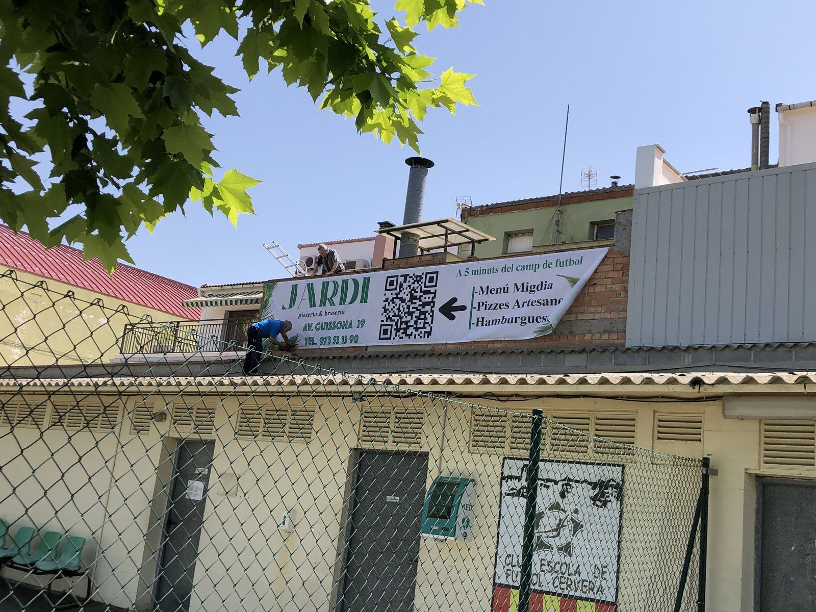 Pancarta del Restaurante Jardí con la carta y un código QR, colgada frente al campo de fútbol de Cervera