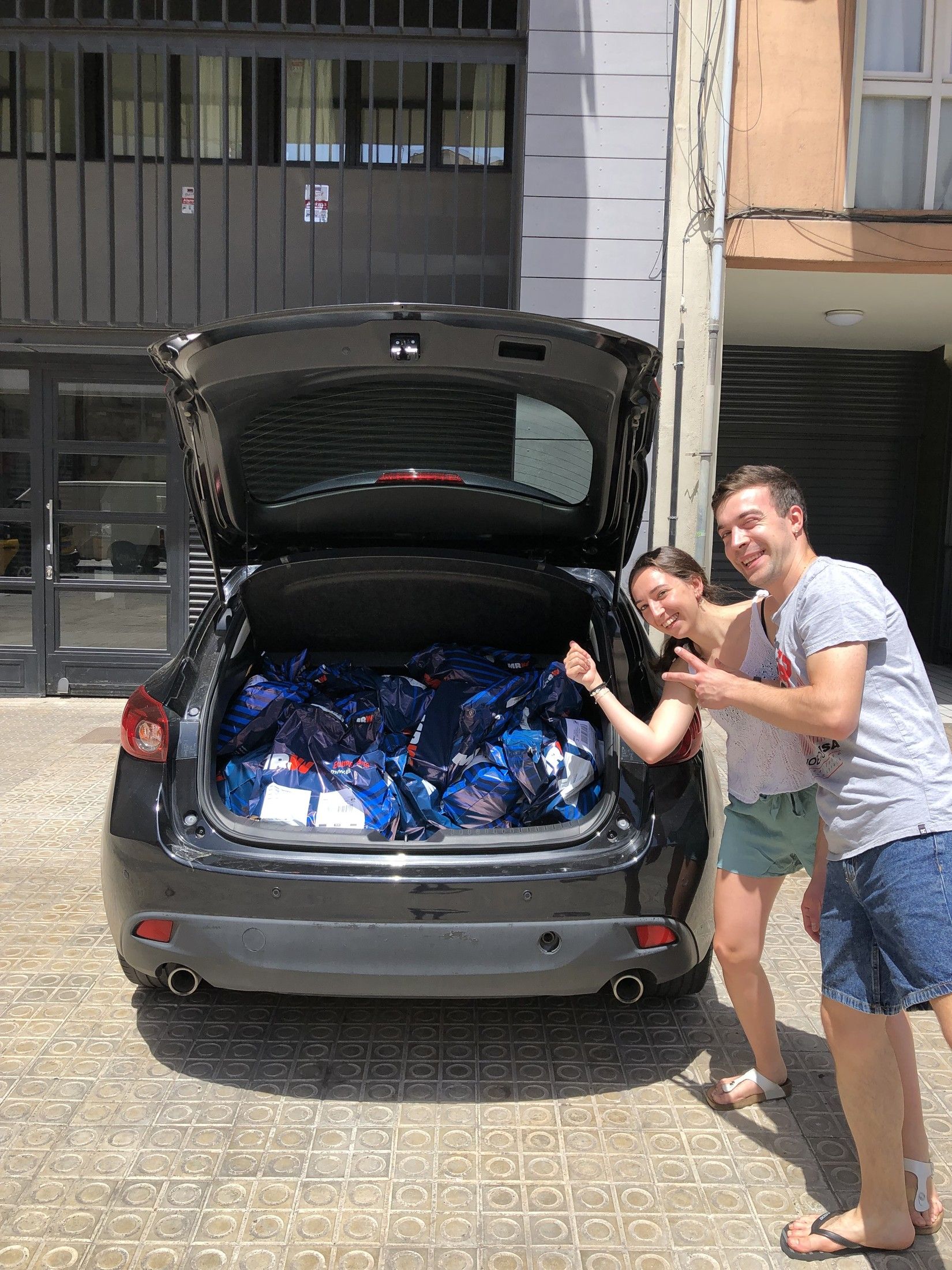 Taras y su compañera de piso sonríen y señalan el maletero abierto de un coche lleno de bolsas azules de MRW con pedidos de LikinShop. Barcelona, junio de 2022.