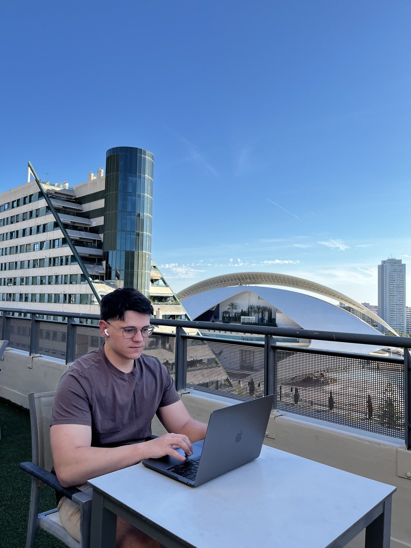 Taras Vasyliv, con gafas y camiseta marrón, trabaja con su portátil en una terraza de Valencia, con los edificios de la Ciutat de les Arts i les Ciències detrás. Mayo de 2024.