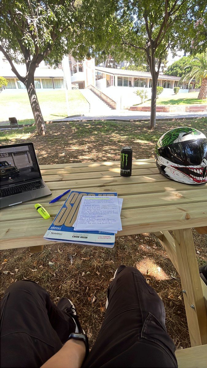Apuntes y un ordenador sobre una mesa de picnic, con el casco de la moto y una lata de bebida energética al lado